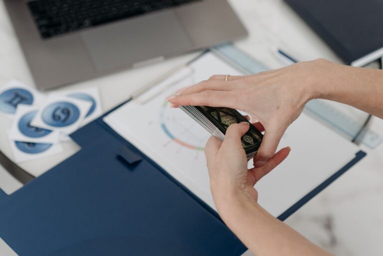 A woman shuffling tarot cards on a desk with a horoscope chart and a laptop.